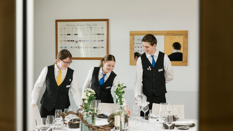 Service in der Tourismusschule Semmering, © Wiener Alpen/Sophie Menegaldo Drei Personen in Uniform decken einen Tisch in einem Schulungsraum ein.