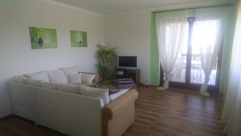 Wohnzimmer, © Anke Spreitzer Ein Wohnzimmer mit einem weißen Sofa, einem kleinen Fernseher, Pflanzen und großen Fenstern mit Vorhängen.