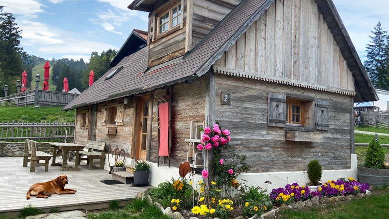 Panoramahütte auf der Mönichkirchner Schwaig, © Wiener Alpen Eine rustikale Holzhütte mit Blumenbeet und einem Hund auf der Terrasse, umgeben von grüner Landschaft und roten Sonnenschirmen.