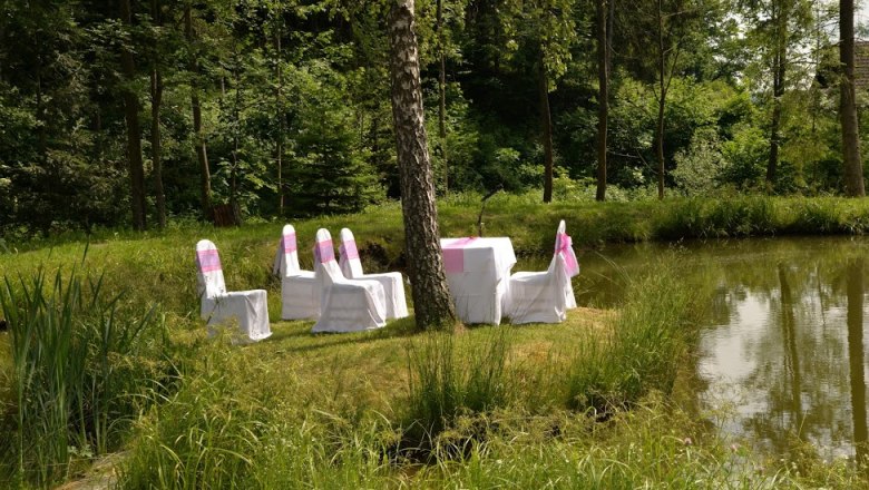 Hochzeit am Teich,, © Hochzeit Gasthaus Buchegger, Foto Buchegger Stühle und ein Tisch mit weißen Hussen auf einer grünen Wiese neben einem Teich, umgeben von Wald.