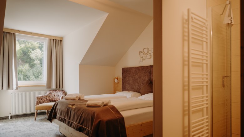 Hotel Post Hönig, © Wiener Alpen, Florian Luckerbauer - Flotoanker Gemütliches Hotelzimmer mit Doppelbett aus hellem Holz, Fenster mit Vorhängen und einem Sessel. Im Vordergrund sieht man ins Badezimmer. Das Zimmer hat fensterseitig eine Dachschräge.