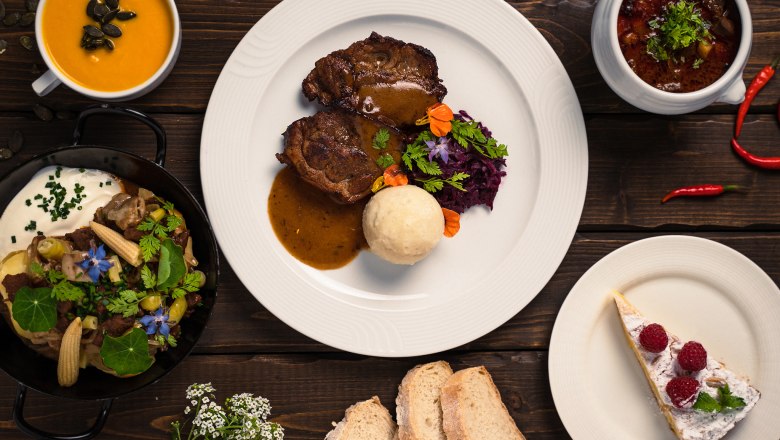 Feine Küche im Sporthotel, © Sporthotel Semmering Verschiedene Gerichte auf einem Holztisch, darunter Suppe, Fleischgericht, Eintopf, Brot und Kuchen.
