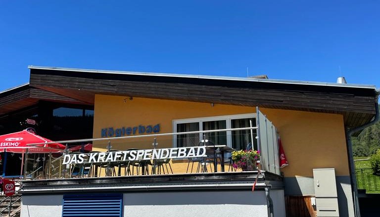 Kögler Bad Rettenegg, © Tourismusverband Oststeiermark, Maria Rein Gelbes Gebäude mit der Aufschrift 'Köglerbad Das Kraftspendebad' und Sonnenschirmen auf der Terrasse.