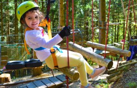 Viel Spaß am Kinderparcours, © Hamari Kletterpark Ein Kind mit Helm und Sicherheitsgurt sitzt auf einem Holzparcours im Wald.