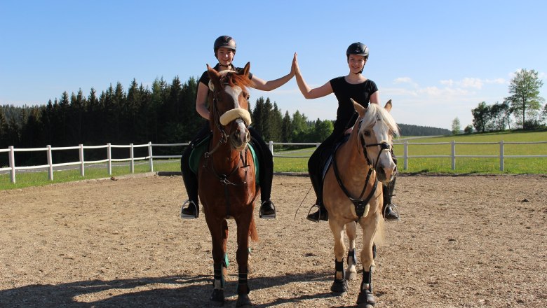 Das Leitner - Narnhoferwirt, © Tourismusverband Oststeiermark, Karin Leitner Zwei Reiterinnen geben sich auf Pferden sitzend ein High-Five auf einem Reitplatz.