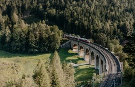 Semmeringbahn, © wieneralpen,nicoleseiser.at Ein Zug fährt über ein Viadukt, Bäume in der Umgebung