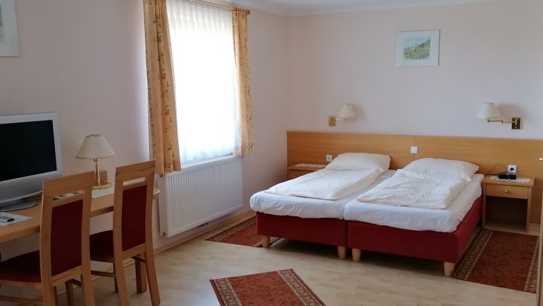 Doppelzimmer, © Wiener Alpen Ein Doppelzimmer mit zwei Einzelbetten, einem Schreibtisch mit Stühlen, einem Fernseher und einem Fenster mit Vorhängen.