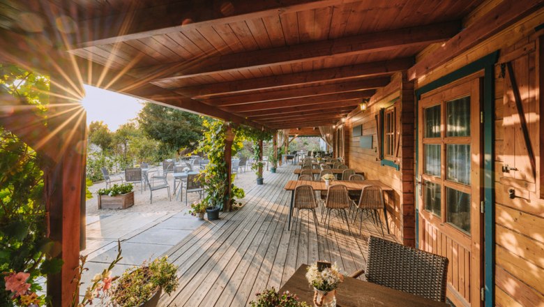 Wunderbare Terrasse zum Genießen - Weinviertler Heuriger, © Martin Mathes Holzterrasse mit Tischen und Stühlen, umgeben von Pflanzen, bei Sonnenuntergang.