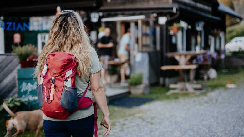 Enzianhütte, © Nixxipixx Fotografie & Design e.U. Nicole Seiser Frau mit rotem Rucksack und Hunde geht auf eine Berghütte zu.