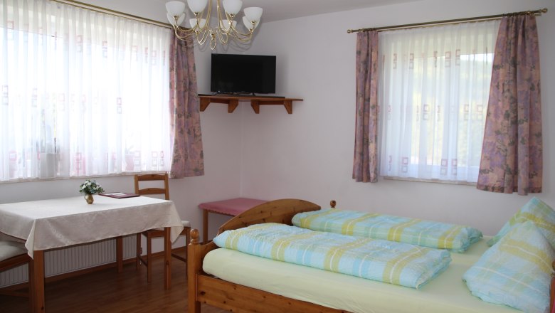 Többágyas szoba, © Karl Buchner Világos szoba franciaággyal, asztallal, székekkel és egy TV-vel a falon.