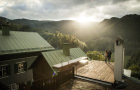 Blick von der Villa, © Matthias Kronfuss Zwei Personen stehen auf einer Terrasse mit Blick auf bewaldete Berge bei Sonnenuntergang.