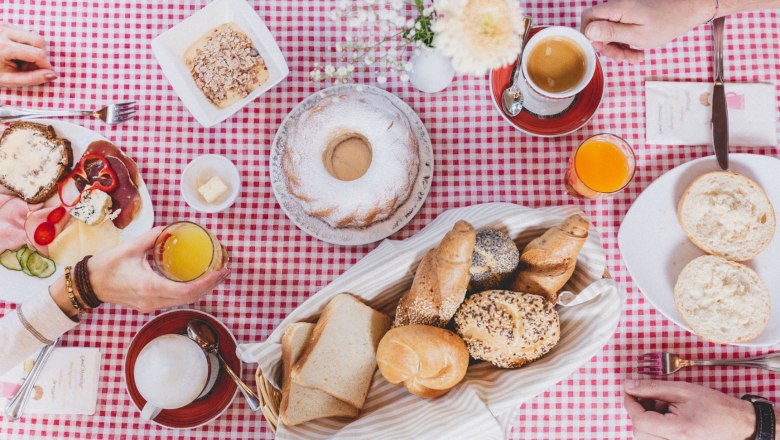 Frühstück, © Gasthof Berghof Frühstückstisch mit Brötchen, Kuchen, Aufschnitt und Getränken auf rot-weiß kariertem Tischtuch.