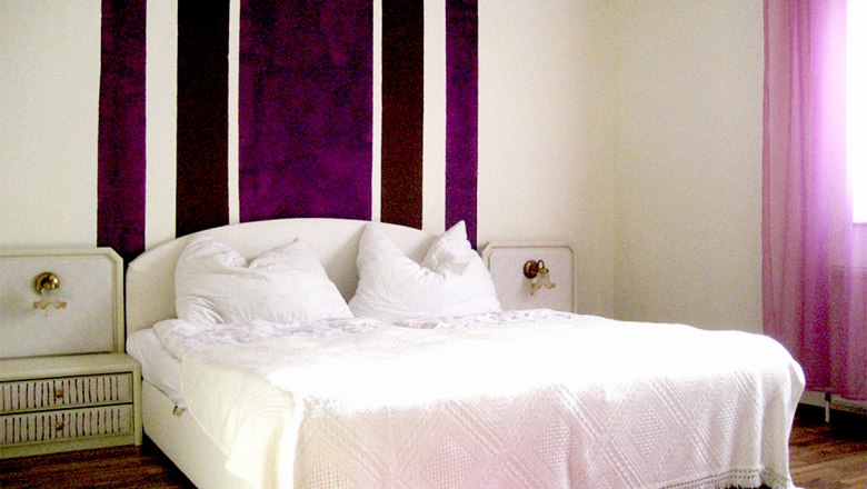 Schlafzimmer 1, © Anke Spreitzer Schlafzimmer mit weißem Bett und lila Streifen an der Wand.