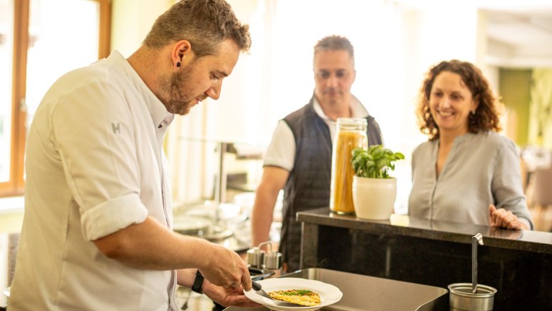 Hotel Mönichwalderhof, © Tourismusverband Oststeiermark, Karl Schrotter Ein Koch legt ein Stück Fisch auf einen weißen Teller. Ein Paar sieht zu.