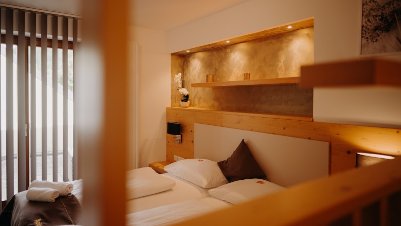 Appartement im Hotel Post Hönig, © Wiener Alpen, Florian Luckerbauer - Flotoanker Gemütliches Appartement mit Doppelbett und Balkontür. Die Wand ist zum Teil mit hellem Holz verkleidet, hinter dem Bett ist eine in die Wand eingelassene Nische mit Ablagefläche und Spots, in einem erdigen Gold gestrichen.