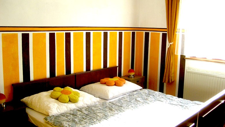 Schlafzimmer 2, © Anke Spreitzer Ein Schlafzimmer mit einem Doppelbett, gestreifter Wand und gelben Vorhängen.