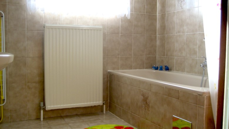 Badezimmer, © Anke Spreitzer Ein Badezimmer mit beiger Fliesenwand, Badewanne, Heizkörper und bunten Teppichen.