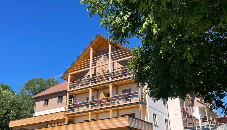 Hotel Schwengerer, © Tourismusverband Oststeiermark, Maria Rein Hotelgebäude mit Holzbalkonen und Carport, umgeben von Bäumen, unter blauem Himmel.