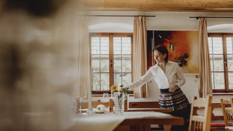 Rundum stimmiges Ambiente, © Niederösterreich Werbung/Sophie Menegaldo Eine Frau in traditioneller Kleidung richtet einen Tisch in einem gemütlichen Raum mit Holzmöbeln und großen Fenstern her.