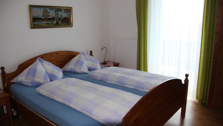 Doppelzimmer, © Karl Buchner Doppelzimmer mit Holzbett, blau-weiß karierter Bettwäsche, Bild an der Wand und grünem Vorhang.