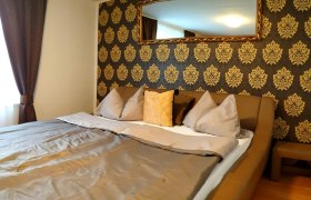 Köszeg Panzió, Zimmer, © Kőszeg-Panzió Ein gemütliches Hotelzimmer mit einem großen Bett, dekoriert mit goldenen Mustern an der Wand und einem Spiegel darüber.