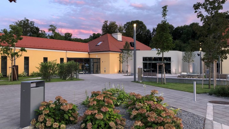 Kulturális színtér Kottingbrunn, © Kulturszene Kottingbrunn Modern épület sárga homlokzattal és piros tetővel, növényekkel és fákkal körülvéve, naplementében.