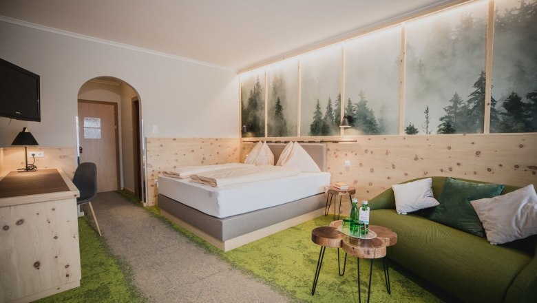 Zirbenzimmer, © Gasthof Berghof Ein gemütliches Hotelzimmer mit Zirbenholzmöbeln, einem Doppelbett, grünem Teppich und einem Sofa. An der Wand ein Bild von nebligem Wald.