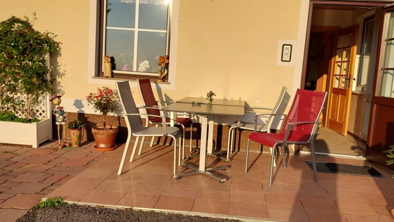 Pension Koderholt, © Koderholt Terrasse mit Tisch und Stühlen vor einem Gebäude, umgeben von Pflanzen.
