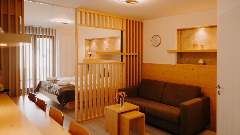 Appartement im Hotel Post Hönig, © Wiener Alpen, Florian Luckerbauer - Flotoanker Modernes Hotelappartement mit Doppelbett, hellen Holzmöbeln, ausziehbarer Couch und Raumtrenner aus Holz. Die Wand ist zum Teil mit Holz verkleidet und in der Wand sind Nischen, die in einem erdigen Gold gestrichen sind.