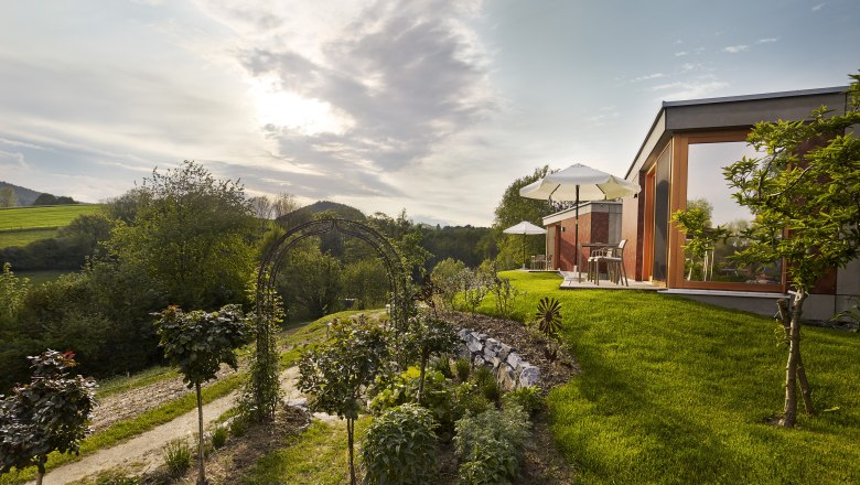 Wiener Alpen Bett: "Bett in da Wies´n", © Bene Croy Moderne Unterkunft in grüner Landschaft mit Sonnenschirmen und Garten.