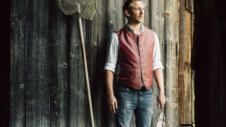 Wirt Sebastian Buchegger, © Niederösterreich Werbung/Julius Hirtzberger Mann in roter Weste steht vor Holzwand mit Fischernetz und Fisch.