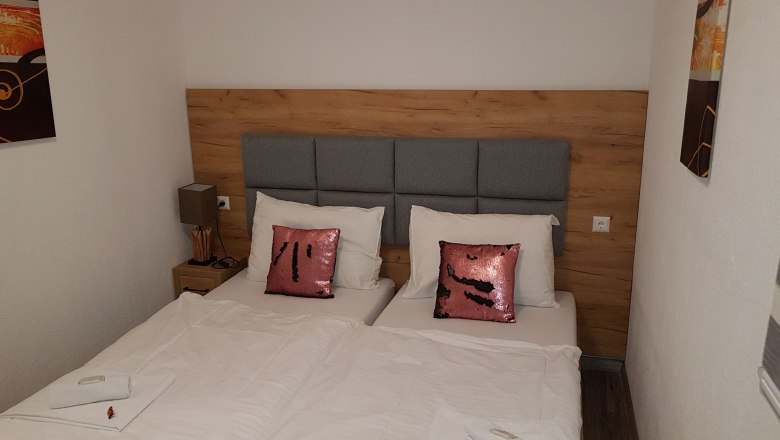 Kétágyas szoba, © Konrad Brandl Kétágyas szoba két egyszemélyes ággyal, szürke fejtámlával, rózsaszín flitteres párnákkal és fehér ágyneművel.