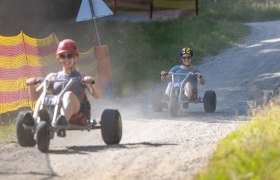 Erlebnisalm Mönichkirchen, © Stefan Wallner Zwei Personen fahren auf Schotterweg mit Mountaincarts, tragen Helme und Sonnenbrillen.
