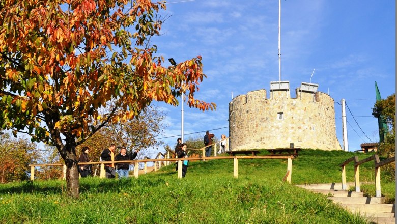 Szuleijmán-Aussichtsturm, © Szulejmán Ein runder Steinturm mit einer Fahne auf einem Hügel, umgeben von grünem Gras und einem Baum mit Herbstlaub.