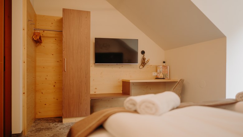 Hotel Post Hönig Zimmer, © Wiener Alpen, Florian Luckerbauer - Flotoanker Ein gemütliches Hotelzimmer fotografiert vom Bett aus. Gegenüber vom Bett hängt ein Flachbildfernsehener an der Wand. Daneben ist eine Garderobe aus hellem Holz, die Wand ist auch zum Teil mit Holz verkleidet. Dort befindet sich auch eine Ablagefläche und ein Schreibtisch mit Sessel und Lampe. Das Zimmer hat eine Dachschräge.