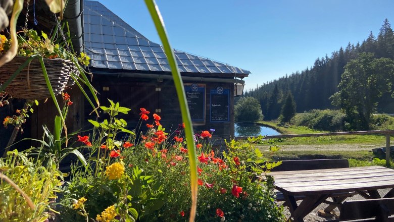 Steyersberger Schwaig, © Wiener Alpen Blumen vor einer Berghütte mit Teich und Wald im Hintergrund.