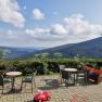 Traumhafter Ausblick auf der Terrasse, © Alpengasthof Fernblick, Fam. Pölzelbauer Terrasse mit Tischen und Stühlen, Blick auf grüne Hügel und blauen Himmel mit Wolken.