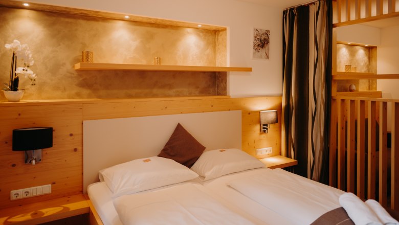 Hotel Post Hönig Appartement, © Wiener Alpen, Florian Luckerbauer - Flotoanker Gemütliches Hotelzimmer mit Doppelbett, heller Holzverkleidung, Raumtrenner aus Holz und mit einem Vorhang. Neben dem Bett sind Steckdosen und Lichtschalter. Über dem Bett eine Nische in der Wand, die beleuchtet und in einem erdigen Goldton gestrichen ist.