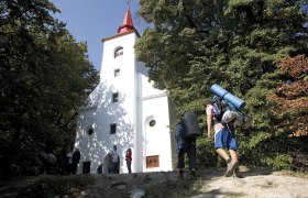 Szent Vid Kapelle, © Szent Vid Wanderer mit Rucksack erreicht eine weiße Kirche im Wald.