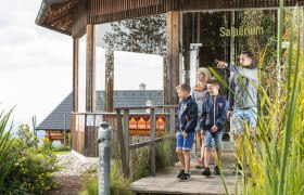 Teil des Themenweges "Lebenswasser", © Erlebnisalm Mönichkirchen, Martin Fülöp Familie auf Holzweg vor einem Gebäude mit der Aufschrift 'Salaerium'.