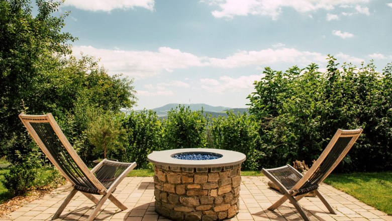 Hotel Schwengerer, © Tourismusverband Oststeiermark, Bernhard Bergmann Zwei Holzstühle und ein Steintisch auf einer Terrasse mit Blick auf grüne Hügel.