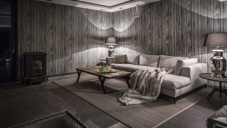 Im Badehaus, © Matthias Kronfuss Gemütlicher Raum mit grauem Sofa, Holztisch, Kamin und Holzvertäfelung.