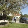 E-Mobilität, © Wiener Alpen Ein Paar geht neben einem geparkten Elektroauto unter einem blühenden Baum in einem ländlichen Garten.