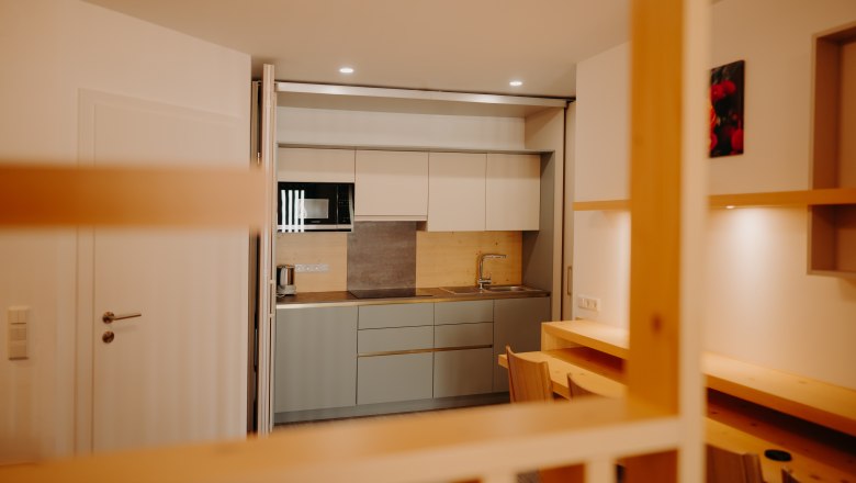 Appartement im Hotel Post Hönig, © Wiener Alpen, Florian Luckerbauer - Flotoanker Moderne Küchenzeile in einem Apartment in hellen Farbtönen. Man sieht eine Spüle, eine Mikrowelle, eine große Herdplatte und einen Wasserkocher. Die Küche kann hinter einer verschiebbaren Trennwand versteckt werden. Vor der Küche sieht man einen Tisch an der Wand und 4 Sessel.