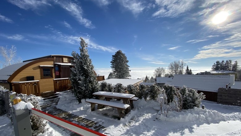 Winterliche Glamping-Hütte mit Schnee bedeckt, umgeben von Bäumen und blauem Himmel.