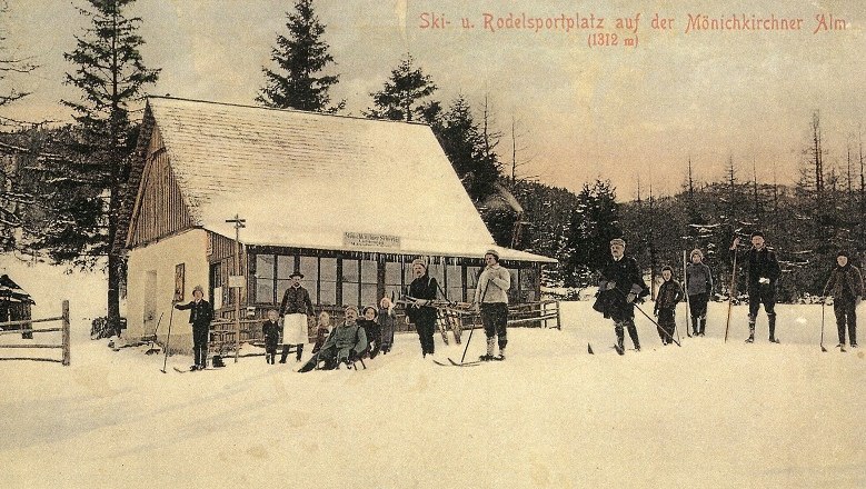 Historische Postkarte eines Skigebiets mit Menschen auf Skiern vor einer Hütte im Schnee.