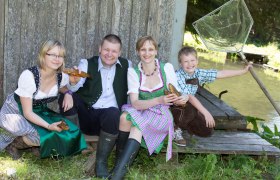 Eine Familie in Tracht sitzt lachend vor einem Teich.