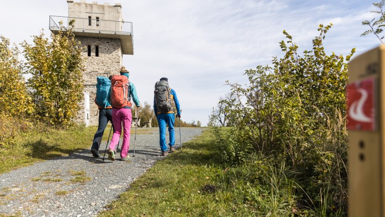 Drei Wanderer mit Rucksäcken gehen auf einem Weg zu einem Steinturm.