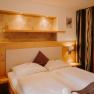 Gem&uuml;tliches Hotelzimmer mit Doppelbett, heller Holzverkleidung, Raumtrenner aus Holz und mit einem Vorhang. Neben dem Bett sind Steckdosen und Lichtschalter. &Uuml;ber dem Bett eine Nische in der Wand, die beleuchtet und in einem erdigen Goldton gestrichen ist.