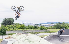 Zwei BMX-Fahrer auf einer Pumptrack-Anlage, einer macht einen R&uuml;ckw&auml;rtssalto, der andere f&auml;hrt eine Kurve.